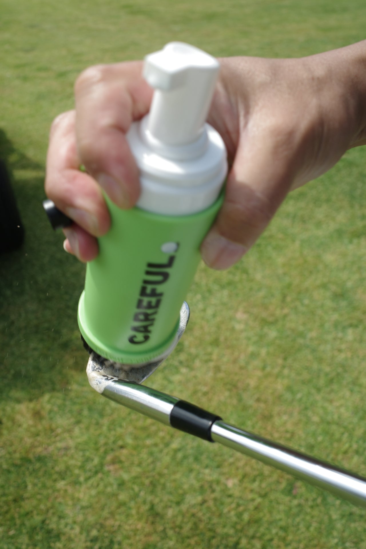 photo of Careful Golf's Patent Pending Caddy Brush with the Brand logo printed on it being held with a hand and cleaning a golf club head on a golf course with the green fairway as the ground/background. 